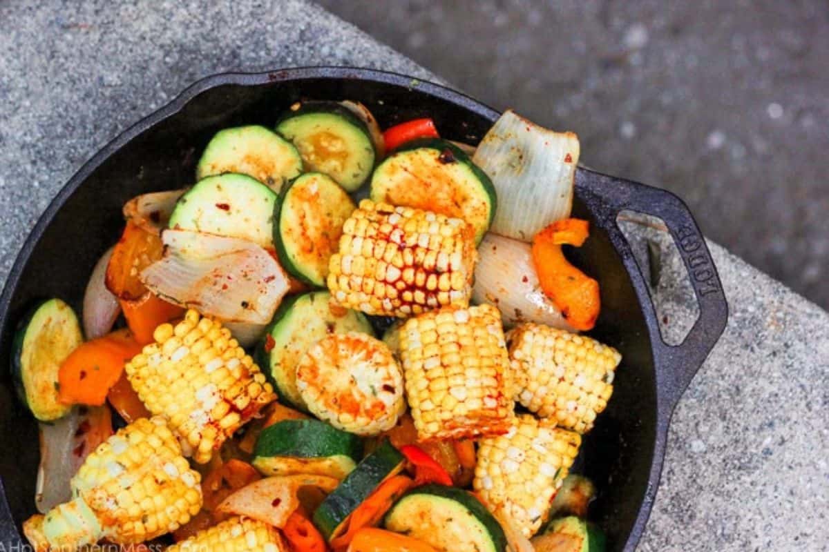 A cast iron skillet filled with flavor-packed grilled corn, zucchini, onions, and bell peppers sits on a gray surface.