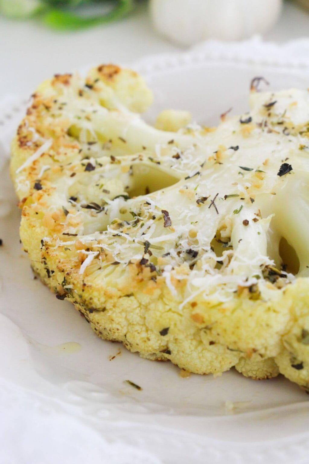 A close-up of Roasted Cauliflower Steaks on a white plate, topped with grated cheese and sprinkled herbs.