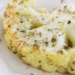 A close-up of Roasted Cauliflower Steaks on a white plate, topped with grated cheese and sprinkled herbs.