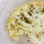 The vegetable steak topped with shredded cheese and herbs sits on a white decorative plate—a delicious way to enjoy Roasted Cauliflower Steaks.