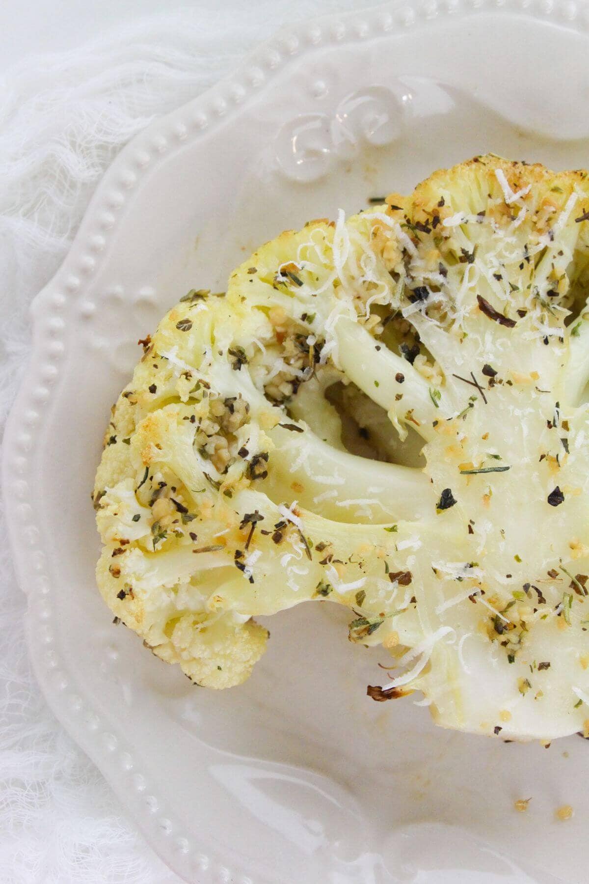 The vegetable steak topped with shredded cheese and herbs sits on a white decorative plate—a delicious way to enjoy Roasted Cauliflower Steaks.