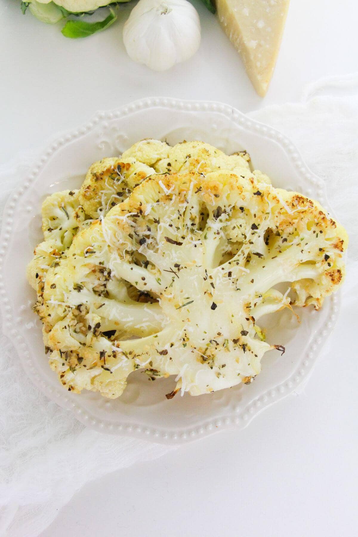 Two of the vegetables garnished with herbs and grated cheese are served on a white plate, with garlic and a wedge of cheese in the background, creating a savory vegetarian dish.
