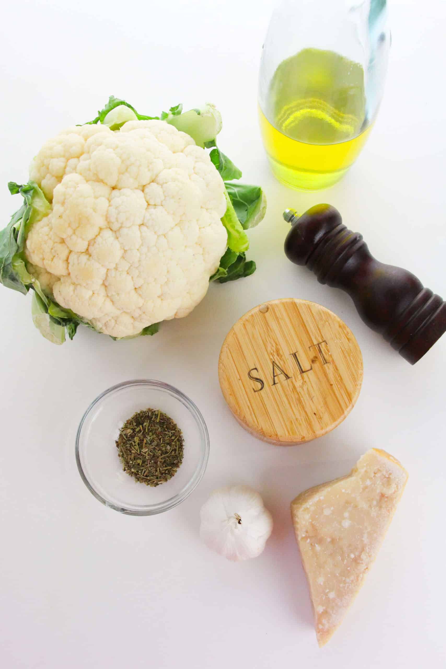 A head of cauliflower, olive oil, a pepper grinder, a salt container, dried herbs, a garlic bulb, and a wedge of Parmesan cheese are arranged on a white surface—perfect for preparing delicious Roasted Cauliflower Steaks.