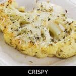 A roasted cauliflower steak, part of our signature Roasted Cauliflower Steaks, is topped with herbs and grated cheese on a white plate, with garlic and a wedge of cheese in the background.