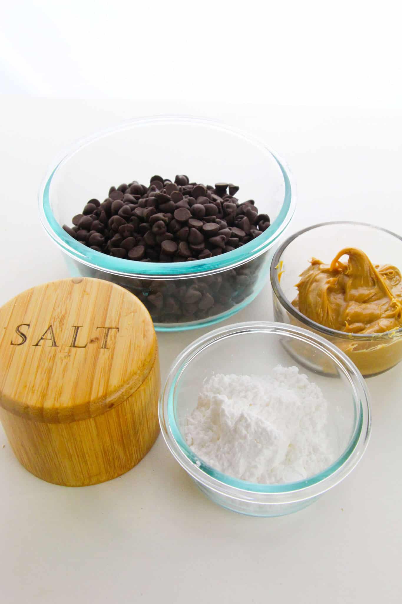 Four ingredients for homemade treat magic rest on a white surface: a bowl of chocolate chips, creamy peanut butter, powdered sugar, and a wooden container labeled "SALT.