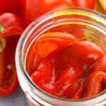 A glass jar filled with vegetables in liquid, surrounded by whole and sliced red bell peppers.