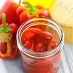 An open jar of roasted red bell peppers in oil sits on a cloth next to whole red bell peppers and a container labeled "salt.