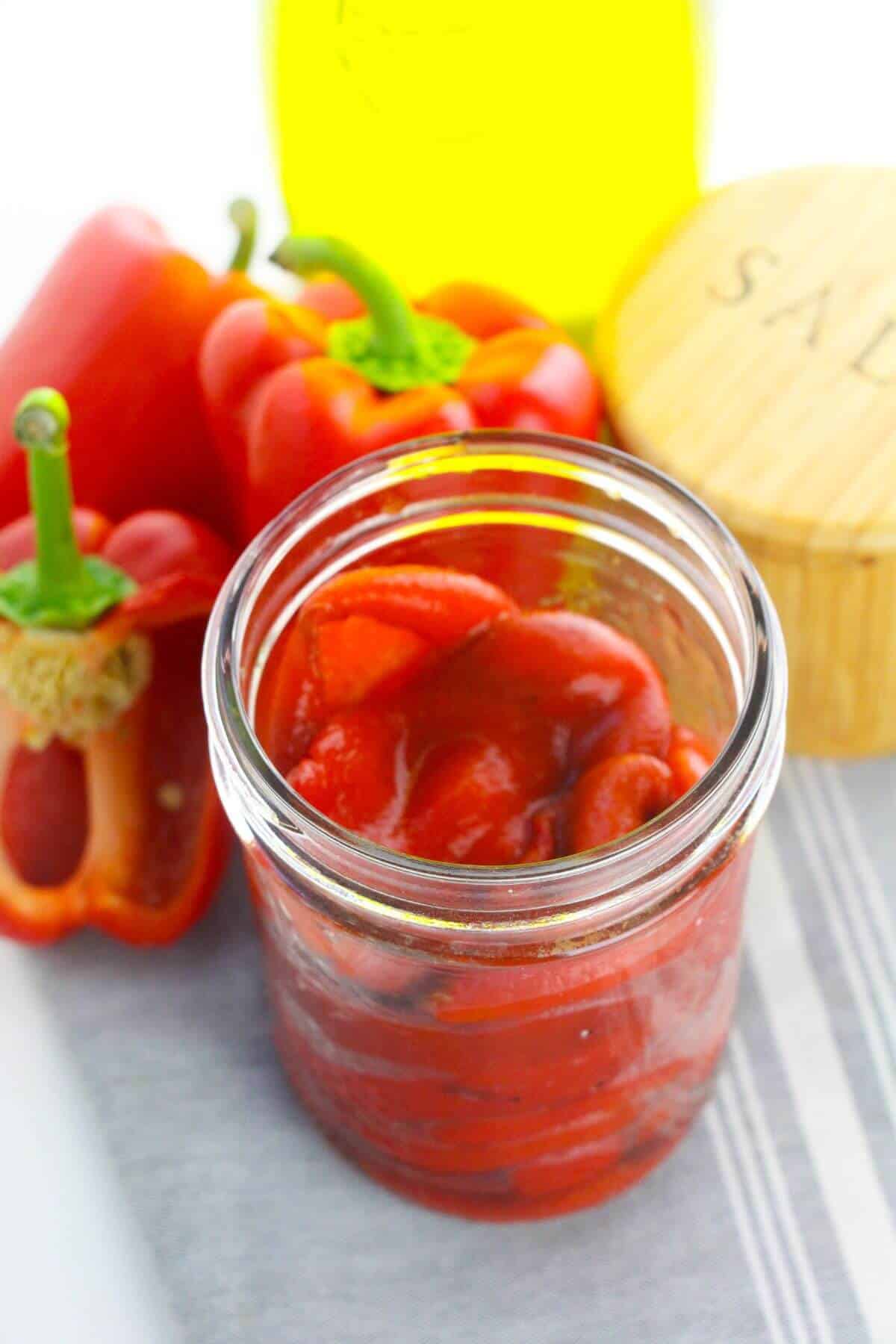 An open jar of roasted red bell peppers in oil sits on a cloth next to whole red bell peppers and a container labeled "salt.