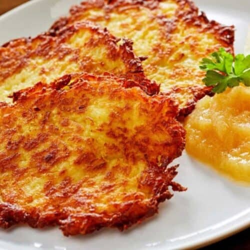 Three crispy potato pancakes on a white plate, served with applesauce, apple slices, and a parsley garnish—one of our German favorites that's perfect for Oktoberfest celebrations.