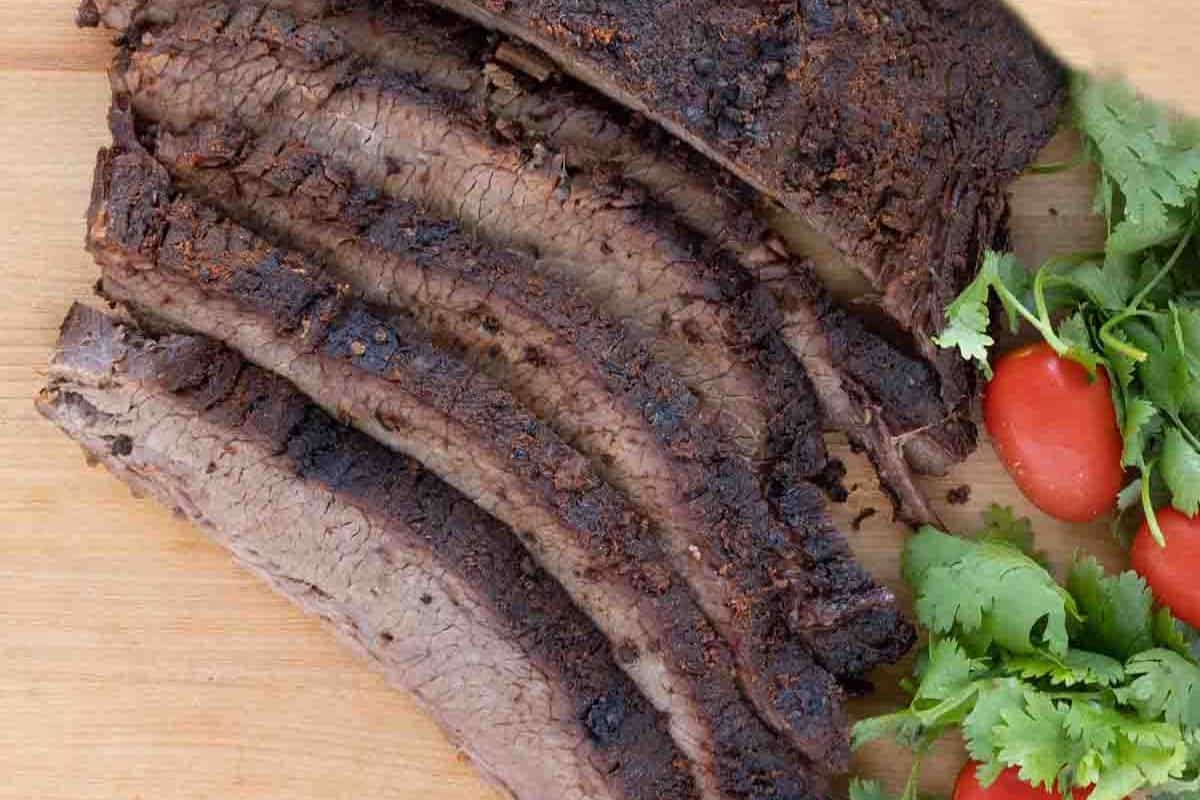 Sliced cooked brisket on a wooden board, with fresh cilantro and grape tomatoes on the side—a true BBQ classic inspired by All American recipes.