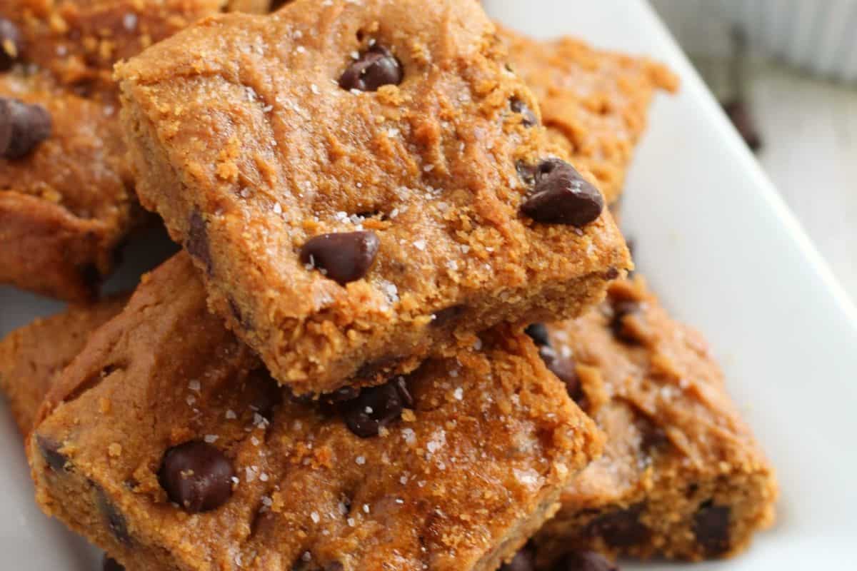 Several chocolate chip blondie bars stacked on a white plate, showing a soft texture and flavor-packed bites.