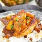 A grilled pineapple pork chop topped with juicy pineapple chunks and sauce is served over a bed of white rice on a decorative plate.