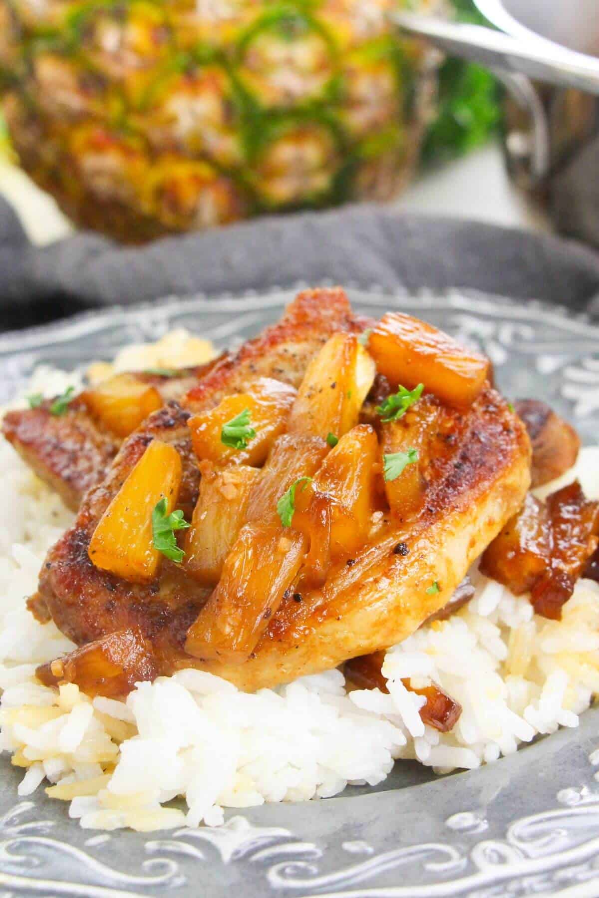 A grilled pineapple pork chop topped with juicy pineapple chunks and sauce is served over a bed of white rice on a decorative plate.