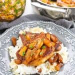 A plate of white rice topped with savory pineapple pork chops and pineapple sauce, garnished with herbs, with a whole pineapple and a pan of food in the background.