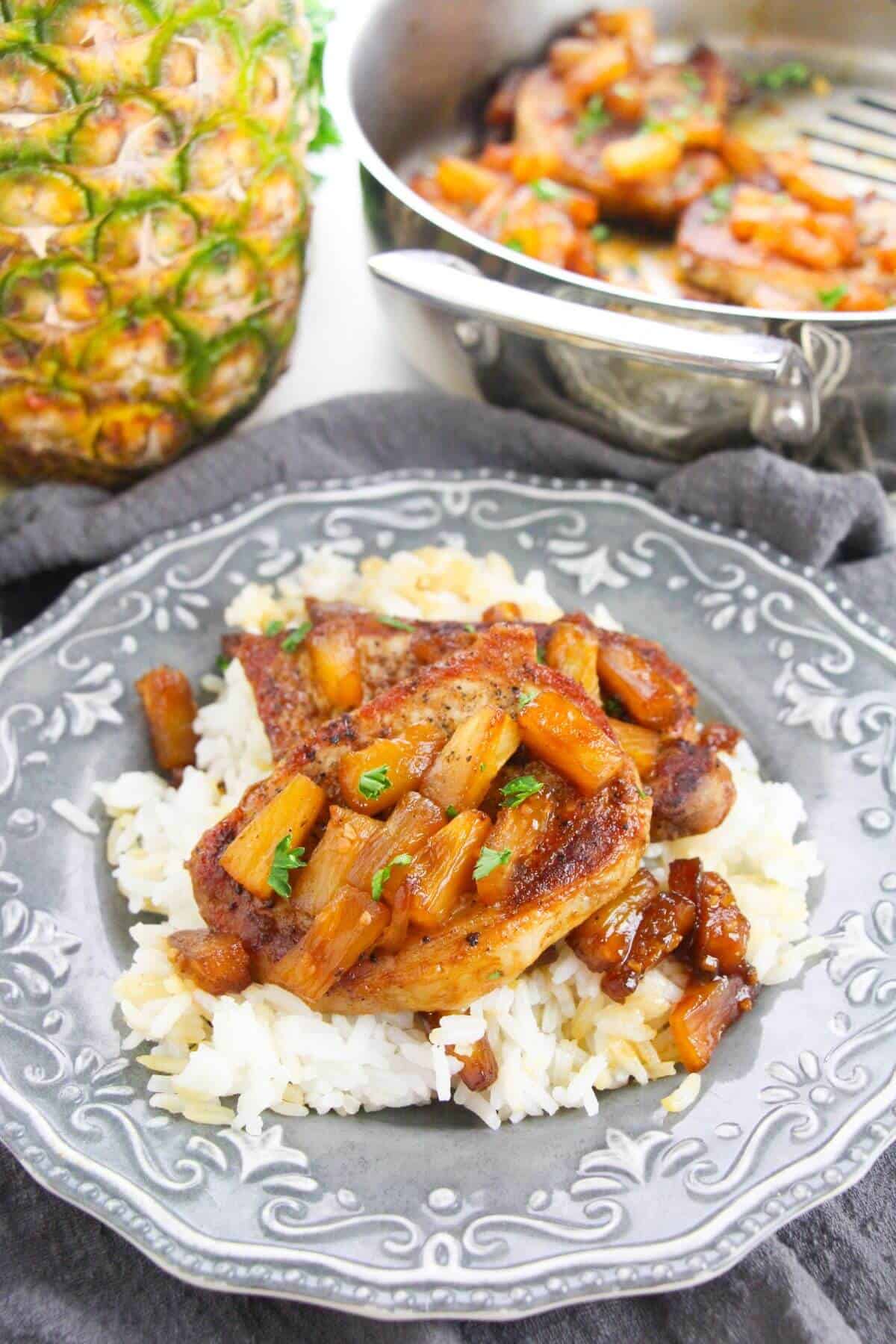 A plate of white rice topped with savory pineapple pork chops and pineapple sauce, garnished with herbs, with a whole pineapple and a pan of food in the background.