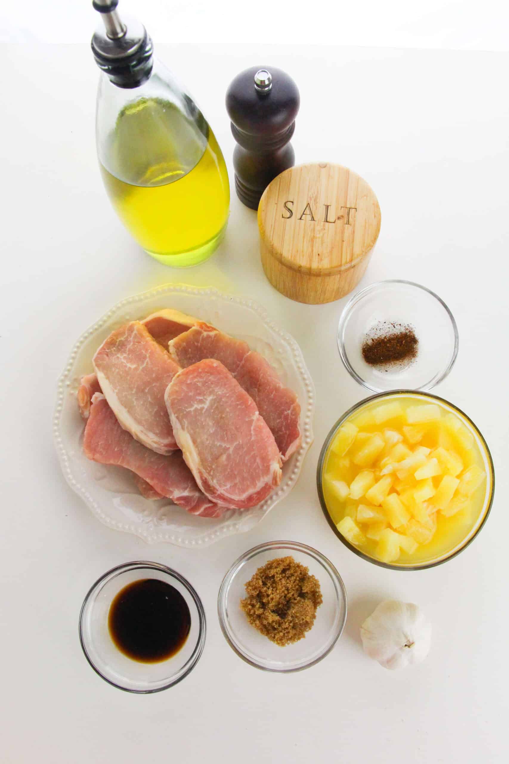 A plate of raw pineapple pork chops is surrounded by a bottle of olive oil, salt, pepper, canned pineapple, garlic, brown sugar, soy sauce, and a small bowl of spices on a white surface.
