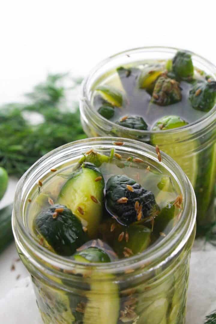 Two glass jars filled with sliced cucumbers in brine, garnished with dill, seeds, and garlic, create the perfect Garlic Dill Pickles, set against a white background.