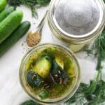An open jar of Garlic Dill Pickles with dill and spices, surrounded by fresh cucumbers, a spoon of seeds, and dill sprigs on a white surface.