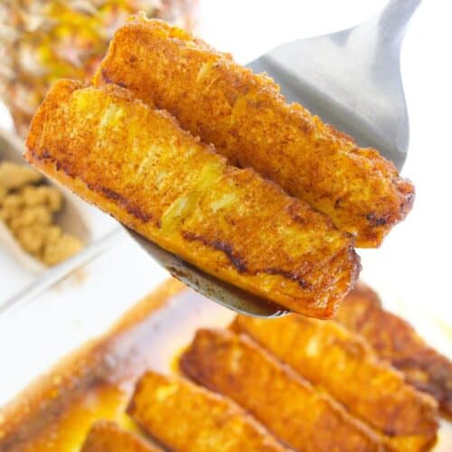 Close-up of a spoon holding roasted pineapple slices caramelized with cinnamon, with more slices and a whole pineapple in the background.