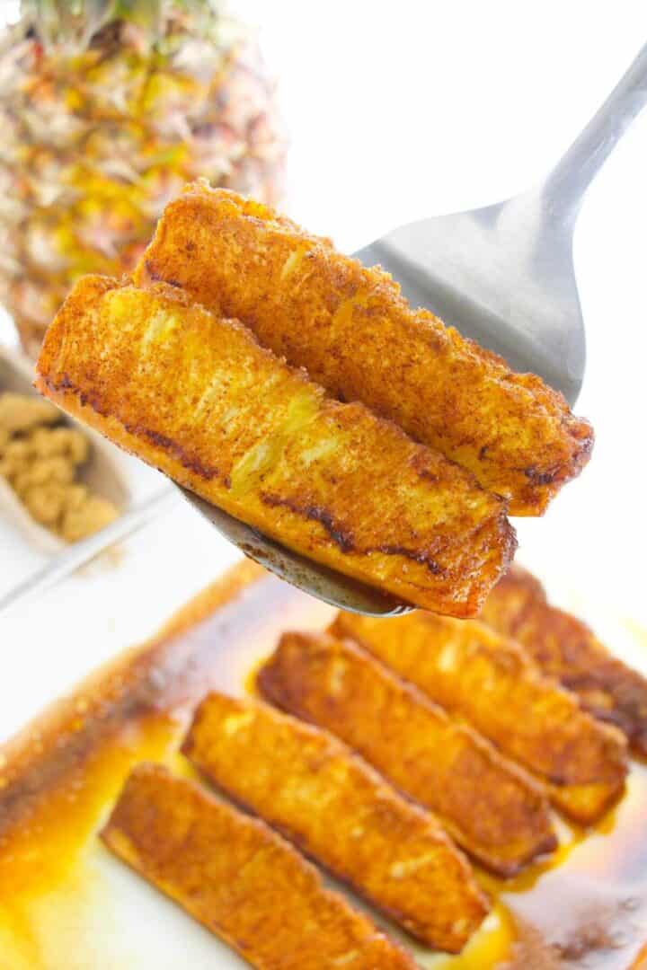 Close-up of a spoon holding roasted pineapple slices caramelized with cinnamon, with more slices and a whole pineapple in the background.