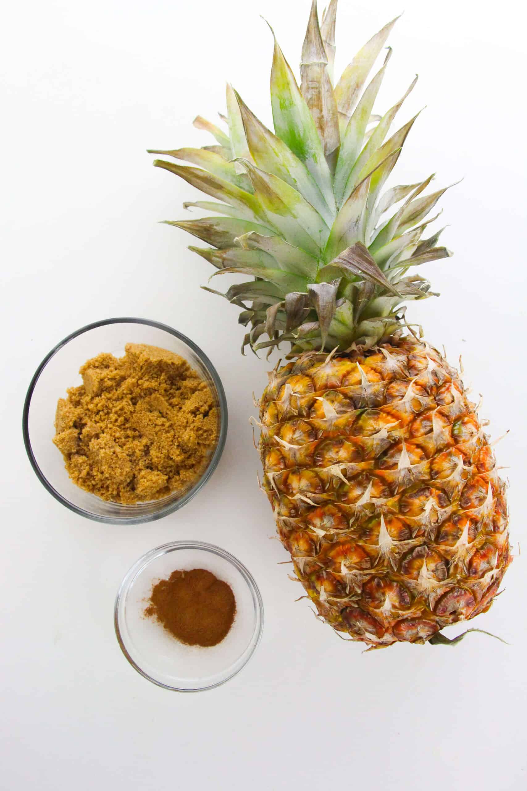 A whole pineapple, a bowl of brown sugar, and a smaller bowl of cinnamon and white sugar are arranged on a white surface.