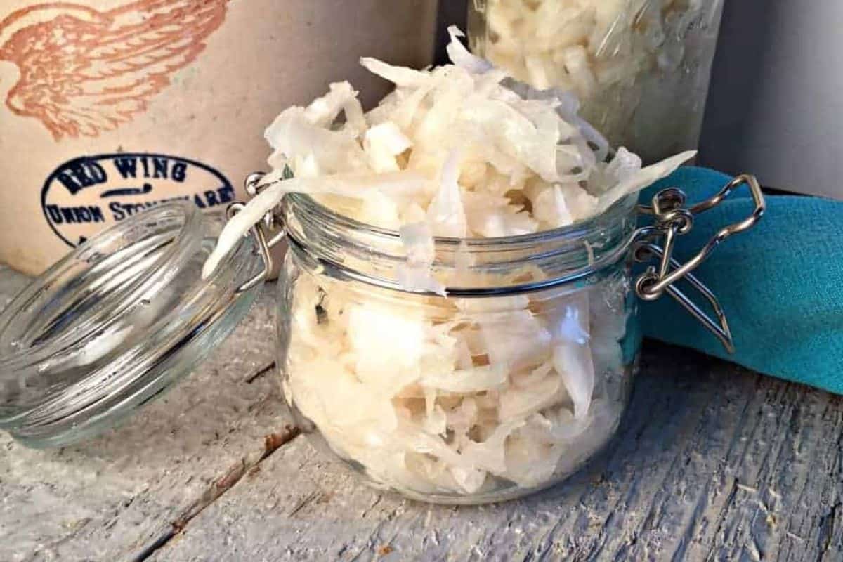 An open glass jar filled with shredded sauerkraut sits on a rustic wooden surface, capturing the essence of German favorites—perfect for celebrating Oktoberfest—with another jar and a crock in the background.