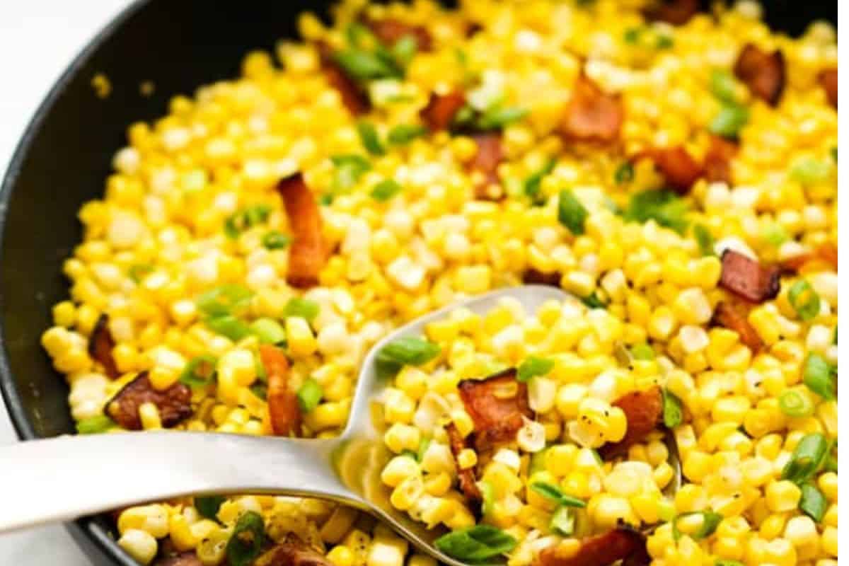 A close-up of a skillet filled with sautéed corn, chopped green onions, and pieces of bacon—an irresistible addition to your favorite summer recipes—with a metal serving spoon resting inside.