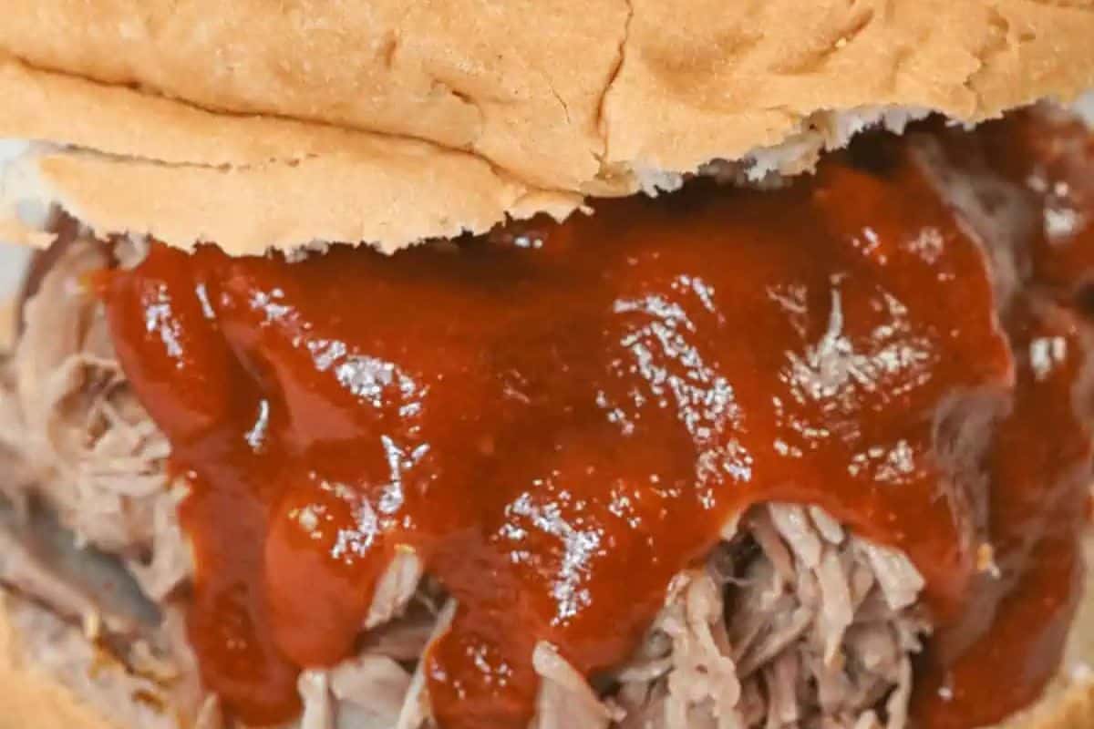A close-up of a Slow Cooker Pulled BBQ Pork sandwich on a bun. Tender, shredded pork is generously coated in a glossy, reddish-brown barbecue sauce.