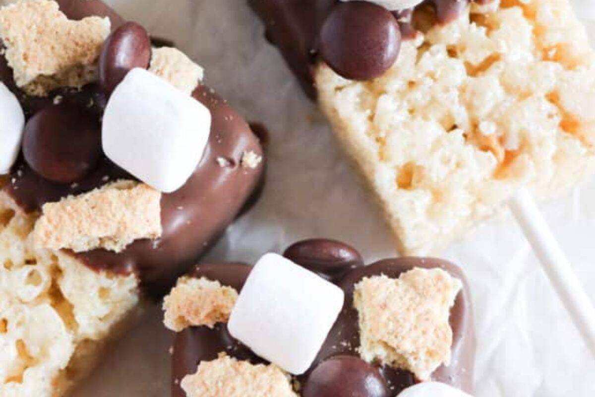 Close-up of flavor packed rice krispie treats on sticks, partially dipped in chocolate and topped with mini marshmallows, graham cracker pieces and chocolate candies.
