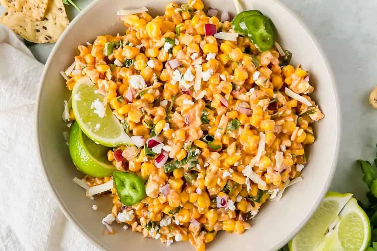 A bowl of Mexican street corn salad, perfect for summer recipes, garnished with lime wedges, jalapeño slices, and shredded cheese sits beautifully on a light surface.