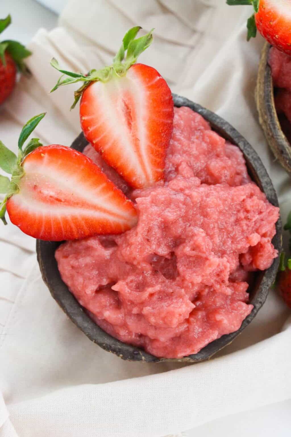 A small bowl of pink strawberry pudding reminiscent of strawberry pudding, is topped with two halved fresh strawberries and placed on a light cloth.