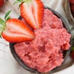 A small bowl of pink strawberry pudding reminiscent of strawberry pudding, is topped with two halved fresh strawberries and placed on a light cloth.