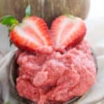 A small bowl filled with pink topped with two halved strawberries; a coconut cup with milk is in the background.