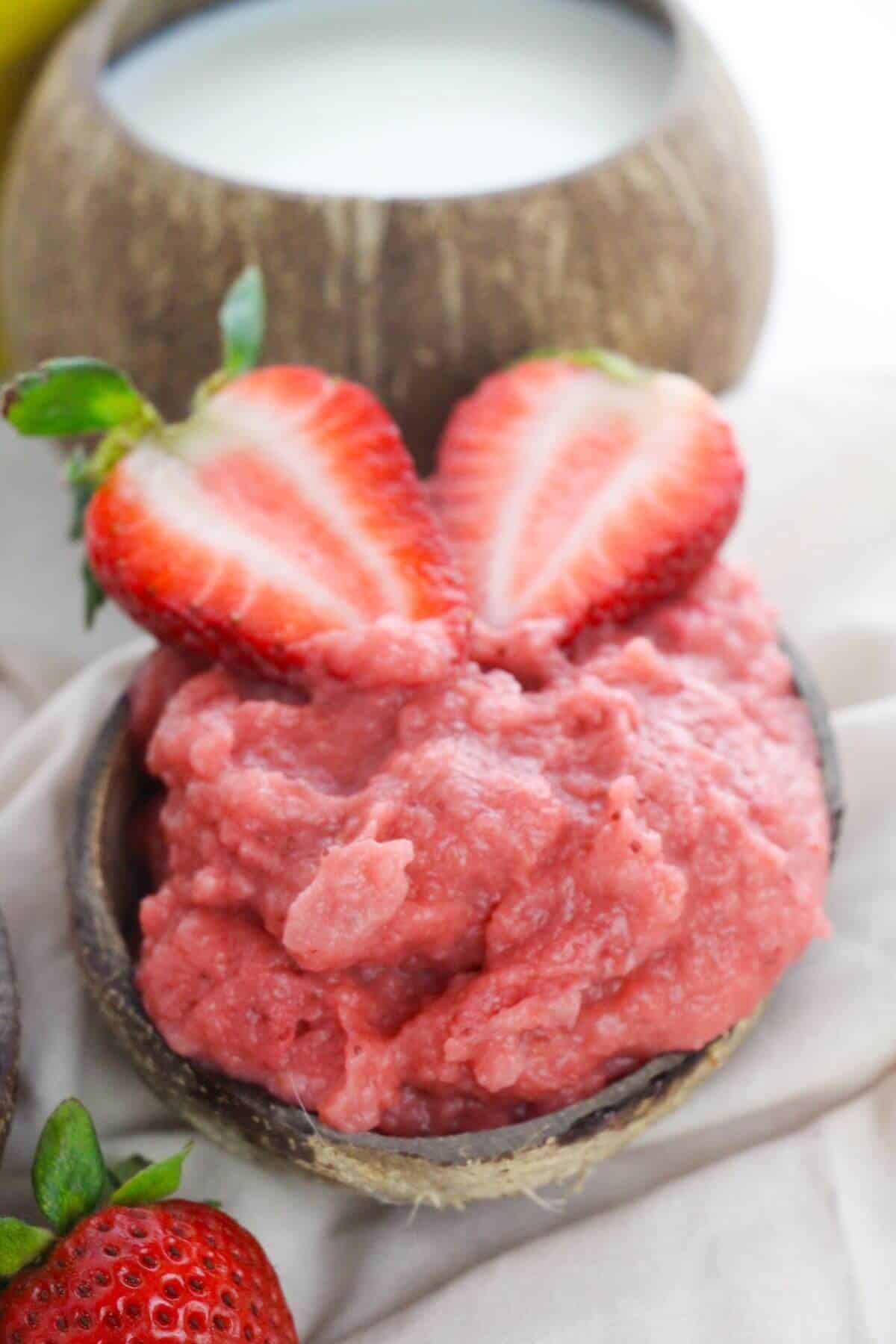A small bowl filled with pink topped with two halved strawberries; a coconut cup with milk is in the background.