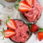 Two bowls of creamy strawberry dessert are garnished with fresh strawberry halves and placed on a light-colored cloth with whole strawberries nearby.