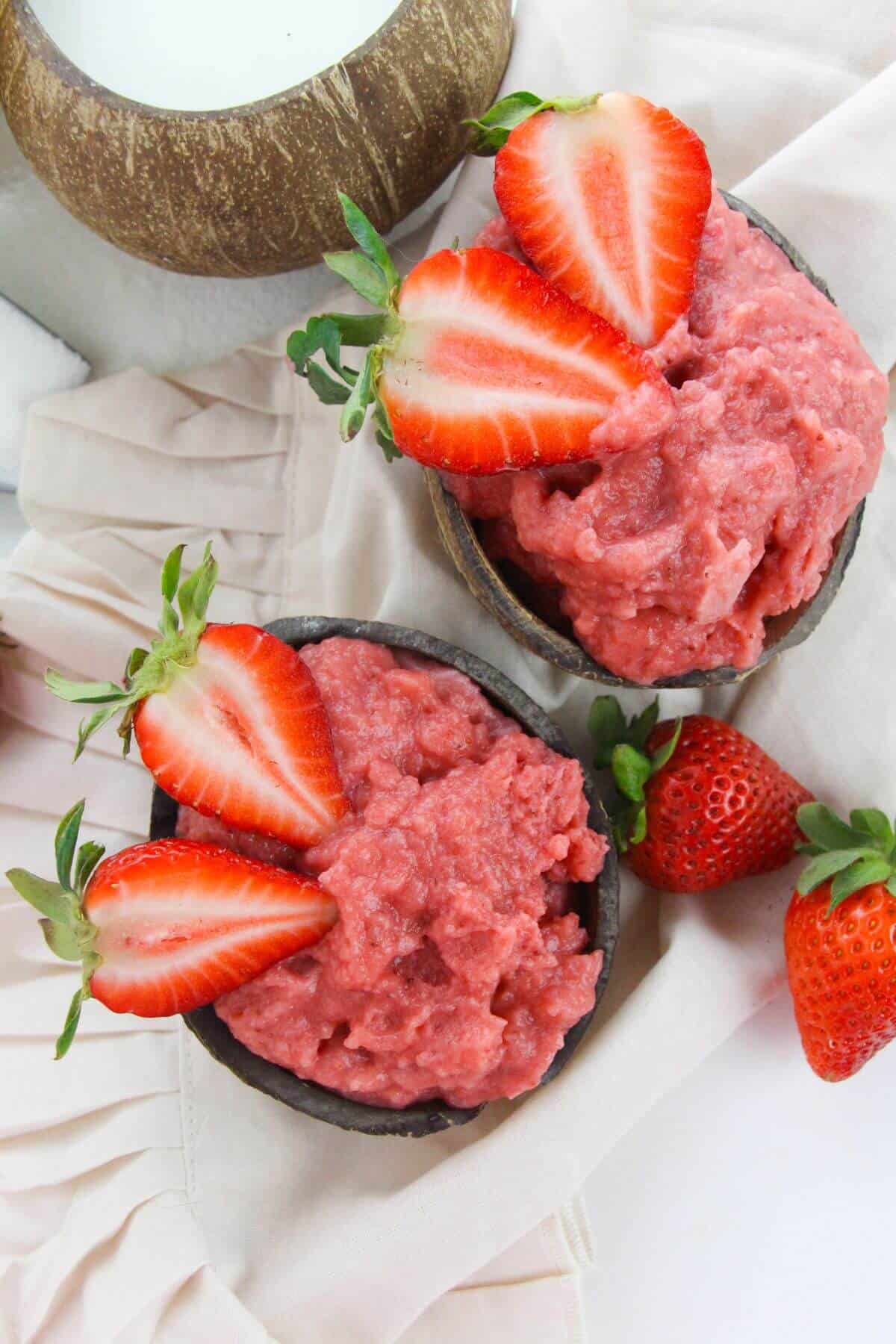 Two bowls  of creamy strawberry dessert are garnished with fresh strawberry halves and placed on a light-colored cloth with whole strawberries nearby.