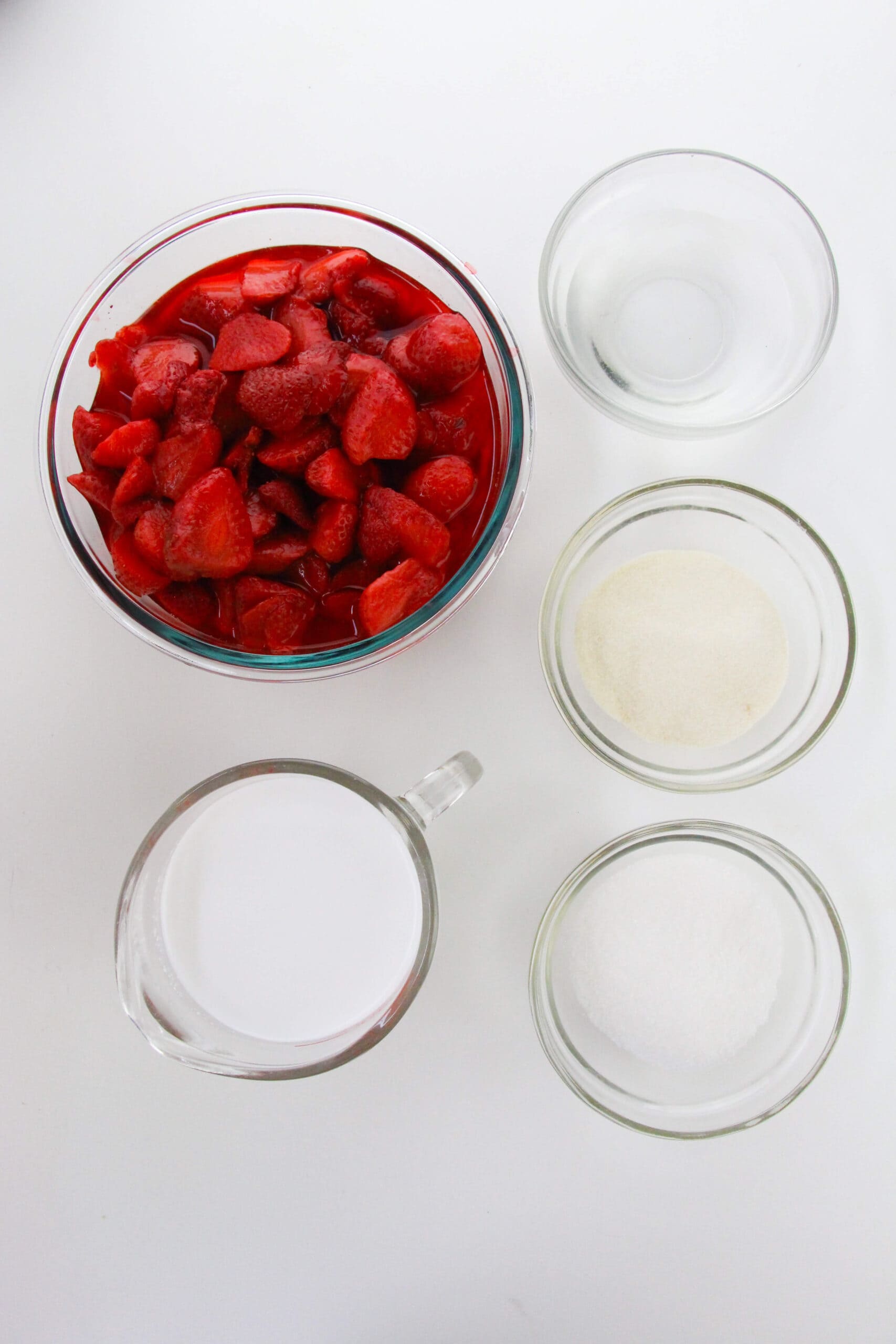 A glass bowl of chopped strawberries in syrup, a measuring cup of milk, and three small bowls with sugar, gelatin, and water are arranged on a white surface—perfect ingredients for homemade dessert.