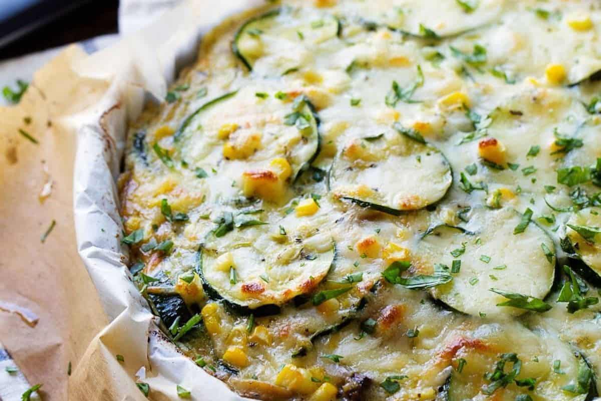 Close-up of a baked vegetable casserole with sliced zucchini, corn on and off the cob, melted cheese, and chopped herbs on top, lined with parchment paper—a perfect addition to your summer recipes.