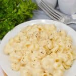 A plate of creamy White Cheddar Mac and Cheese topped with black pepper sits on a wooden board next to fresh parsley, a fork, and salt and pepper shakers.