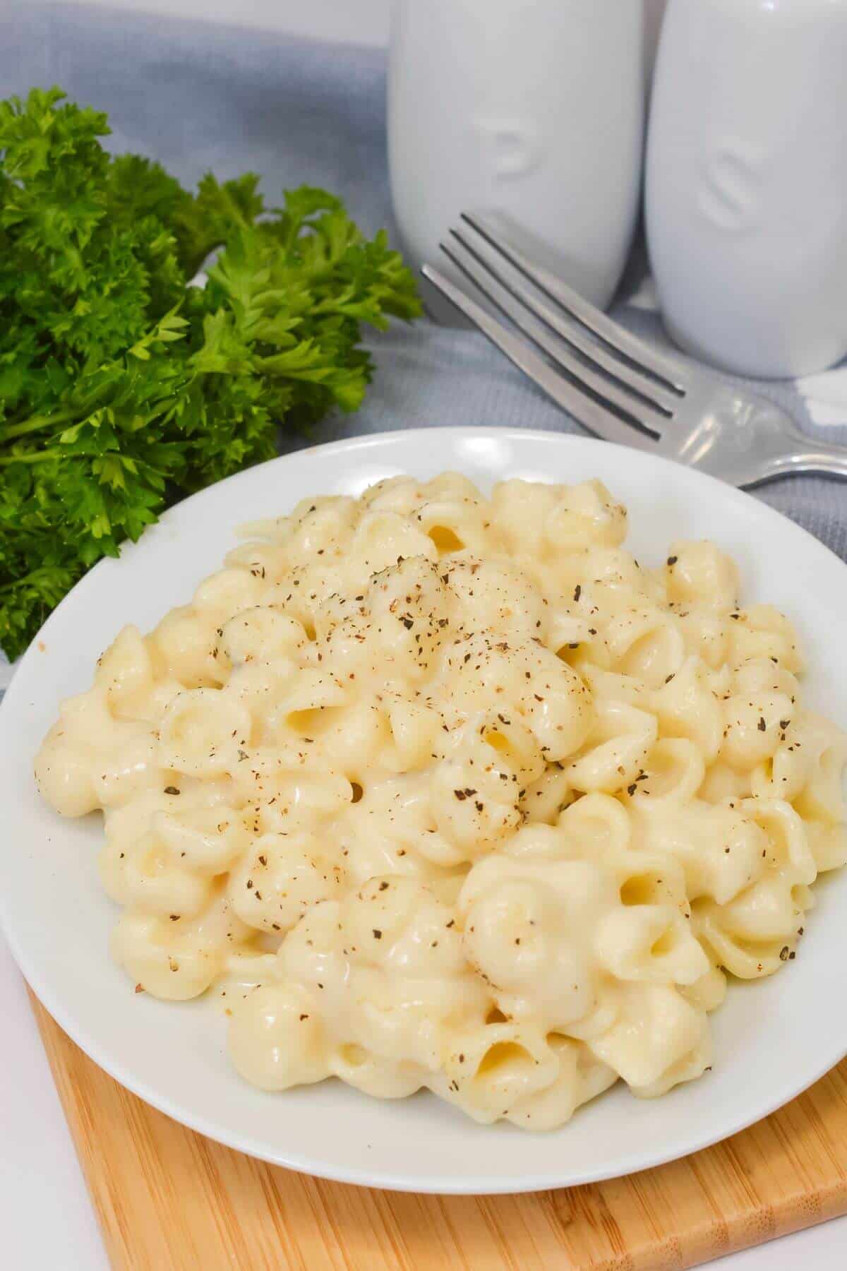 A plate of creamy White Cheddar Mac and Cheese topped with black pepper sits on a wooden board next to fresh parsley, a fork, and salt and pepper shakers.