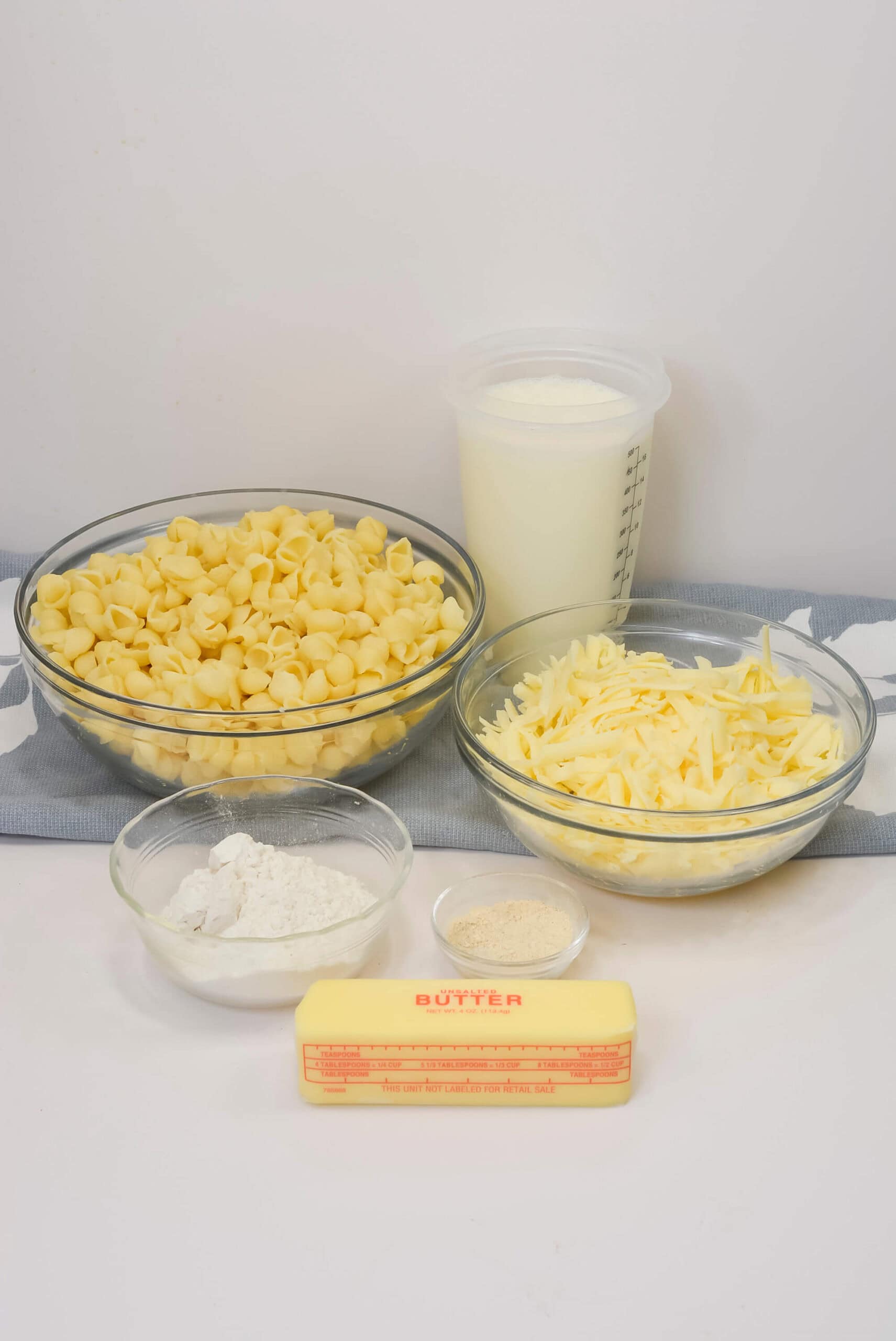 A stick of butter, a bowl of uncooked pasta shells, shredded a bowl of flour, seasoning, and a container of milk rest on a blue kitchen towel.
