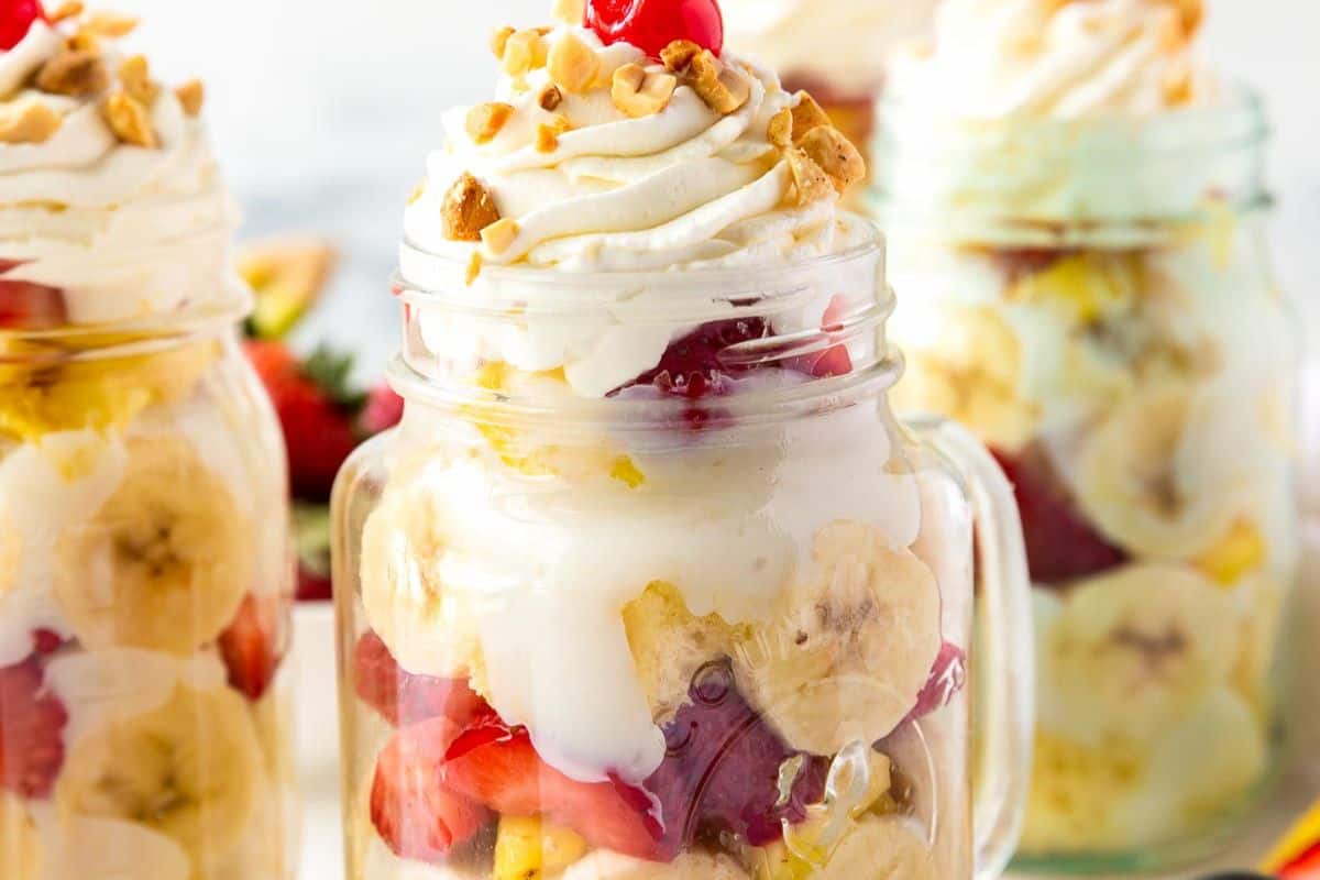 Glass jars filled with layered banana slices, strawberries, pineapple, whipped cream, chopped nuts, and a cherry on top—one of those wow desserts perfect for your collection of easy dessert recipes.