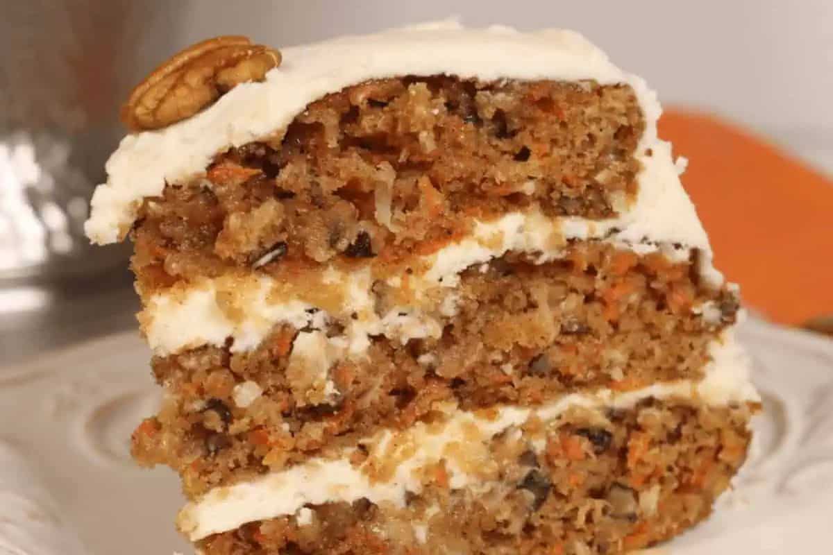 A slice of layered carrot cake with cream cheese frosting and a pecan on top, served on a white plate—perfect for anyone who loves coconut desserts.