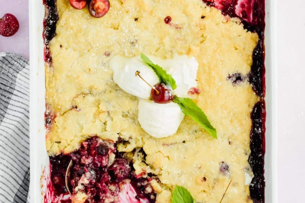 A baked cherry cobbler in a white dish, made with fresh cherries, topped with whipped cream, cherries, and mint leaves, with a portion scooped out—perfect for those who love fresh cherry recipes.