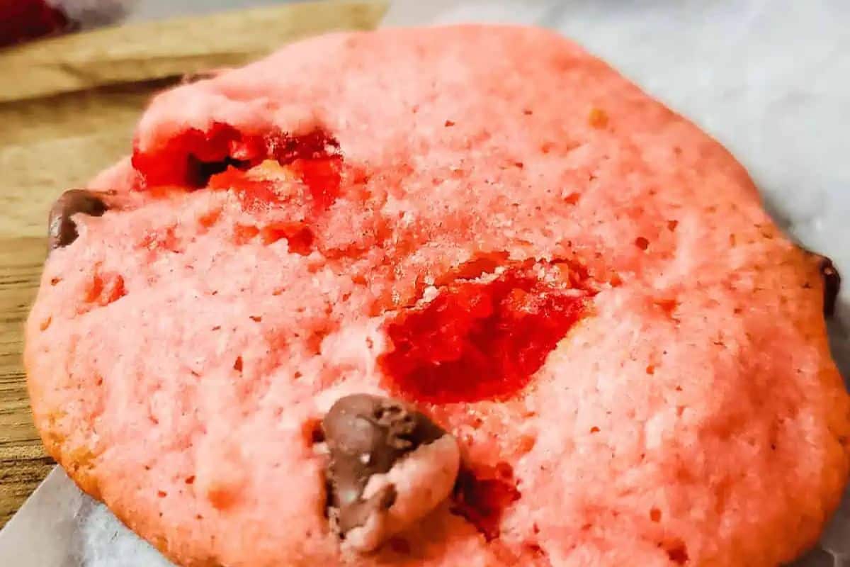 A close-up of a pink cookie with chocolate chips and visible red candied cherry pieces on a wooden and parchment paper surface—perfect for anyone seeking unique cherry desserts or fresh cherry recipes.