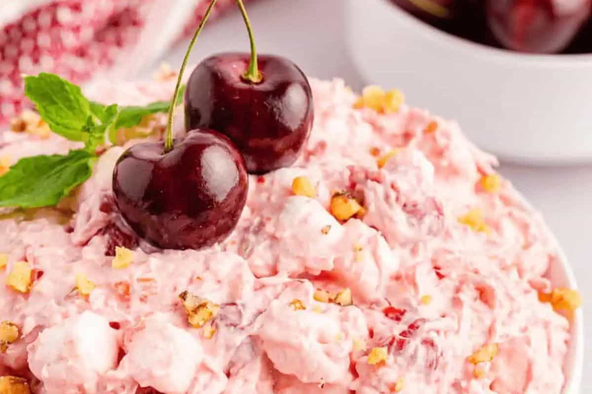 Close-up of a creamy pink fruit salad topped with two fresh cherries, chopped nuts, and a mint sprig—an irresistible treat perfect for lovers of fresh cherry recipes, with more cherries and salad visible in the background.