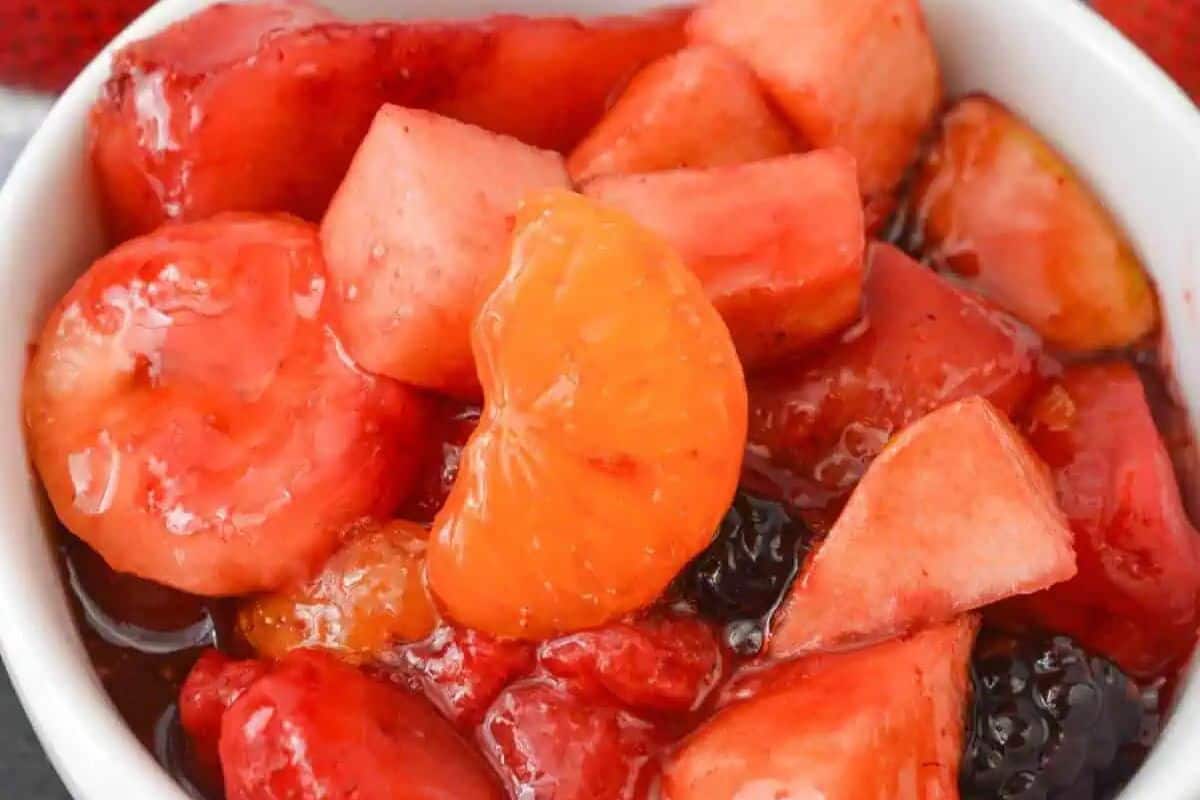 A close-up of a bowl filled with mixed fruit pieces, including mandarin orange segments, apple chunks, and cherries inspired by Fresh Cherry Recipes, all coated in a syrupy sauce.