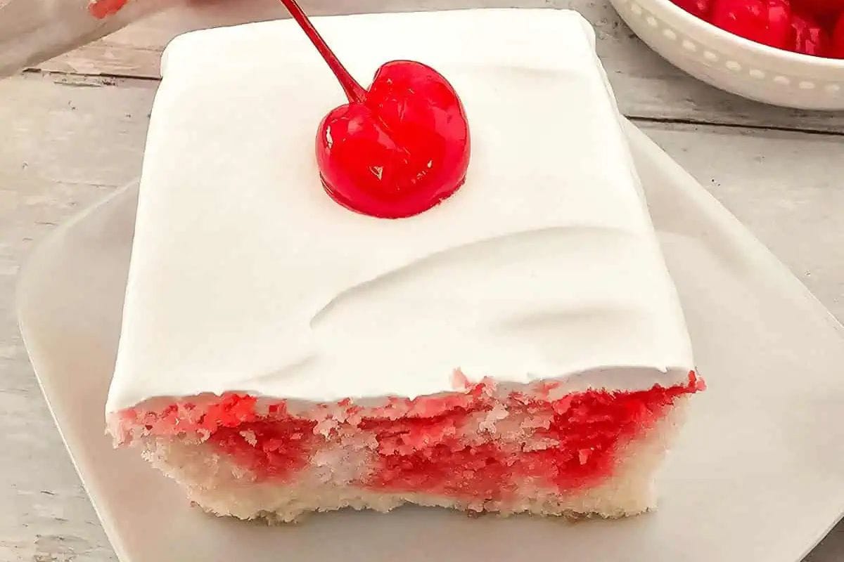 A slice of white sheet cake with red filling and white frosting, topped with a maraschino cherry, on a white plate—perfect for fans of cherry desserts.