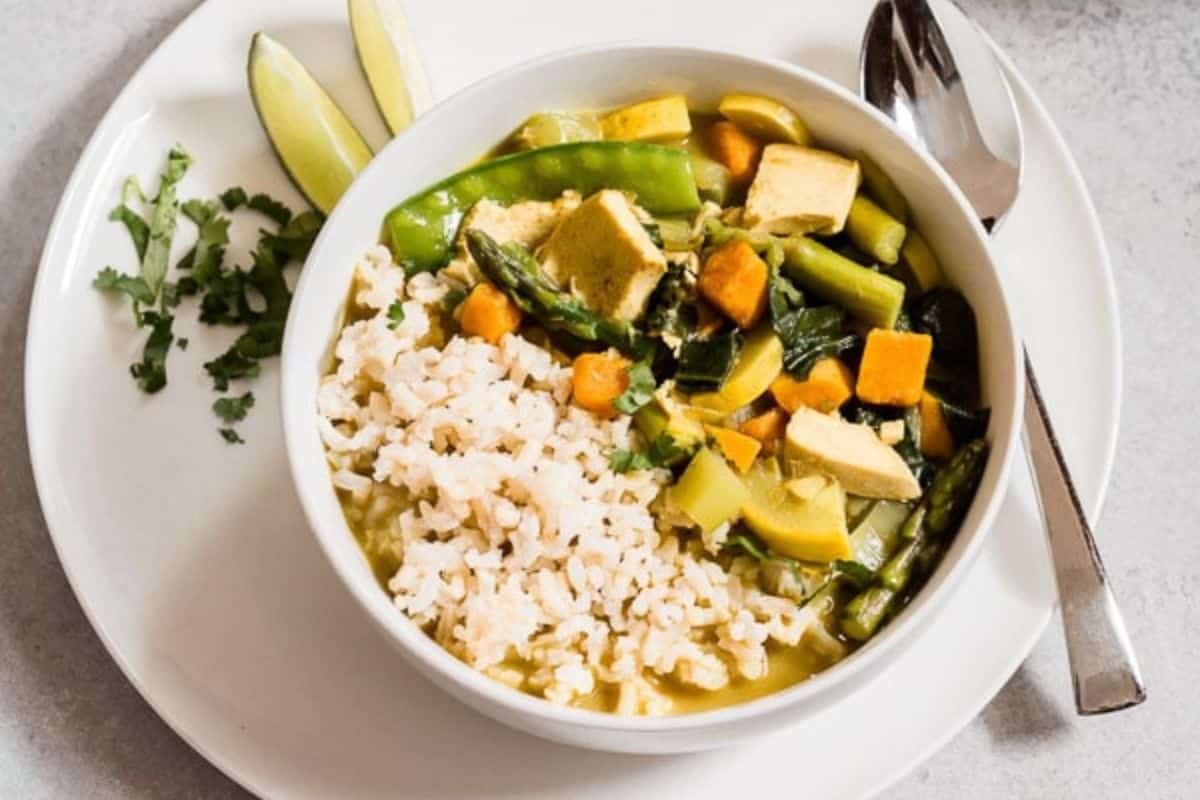 A bowl of brown rice with mixed vegetables and tofu in fragrant coconut curry sauce, served with lime wedges and cilantro on a white plate with a spoon and fork—one of our irresistible coconut recipes.