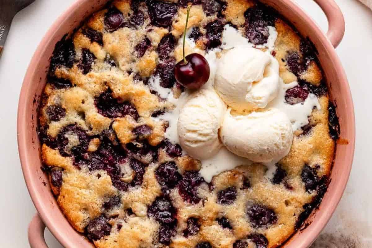 A baked cherry cobbler in a round dish topped with three scoops of vanilla ice cream and a single cherry, perfect for showcasing fresh cherries in delicious cherry recipes.